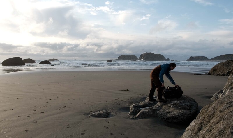 Photographer bending over camera equipment on a large rock at the water's edge of a sandy beach with sea stacks visible offshore.
