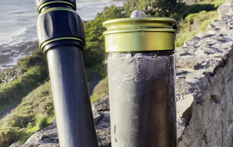 Two cylindrical metal containers with brass bands positioned on rocky coastal terrain overlooking water.