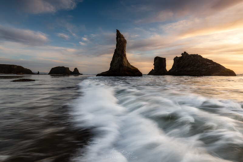 Dramatic rocky sea stacks rise from turbulent waves at sunset with golden light breaking through clouds.
