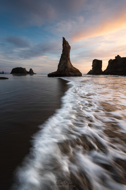 Dramatic coastal rock formation rising from the ocean at sunset, with waves rolling onto a dark sand beach.