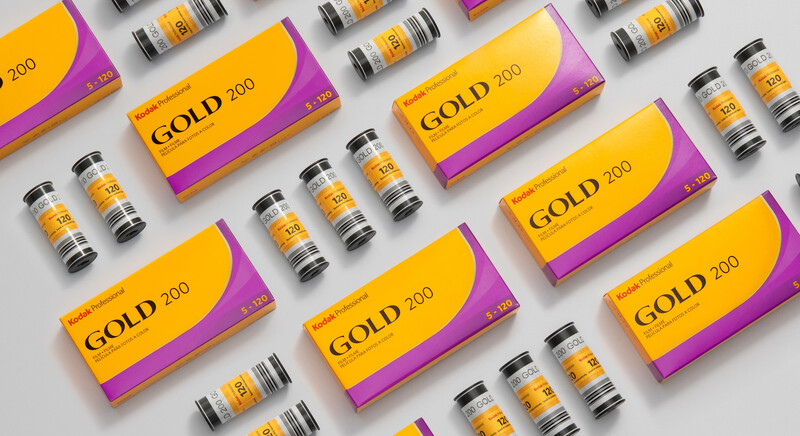 Flat lay arrangement of gold and purple product packaging boxes and cylindrical bottles on a light gray surface.
