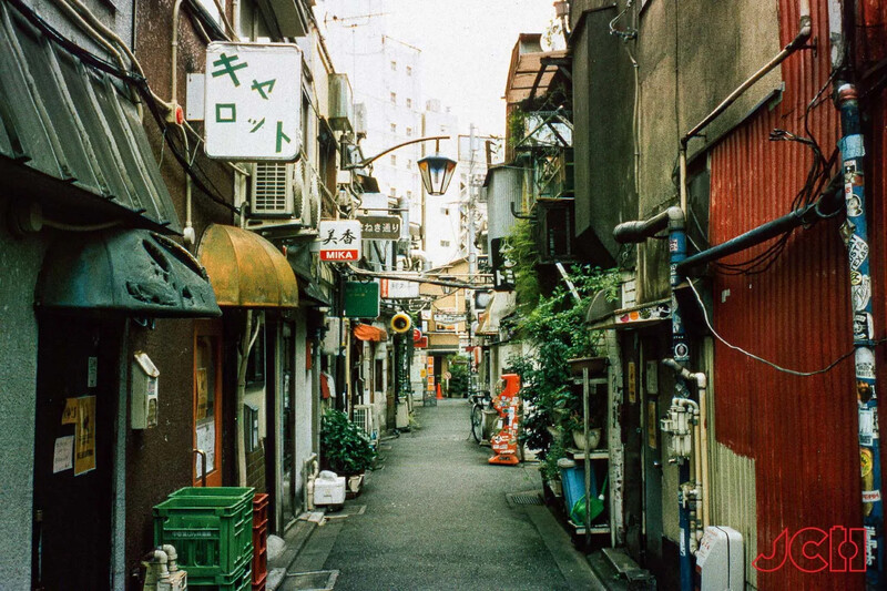 Narrow alley lined with weathered buildings, shops, and vegetation in an urban residential area.