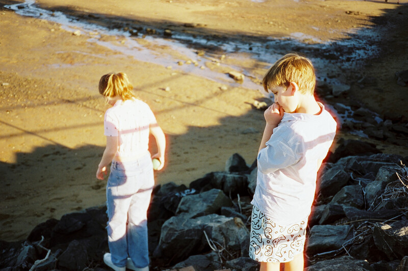 Two young children in white shirts standing on dark rocks beside a sandy beach with shallow water.