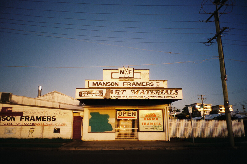 Weathered storefront with stacked vintage signs reading "Manson Framers" and "Art Materials" against twilight sky.