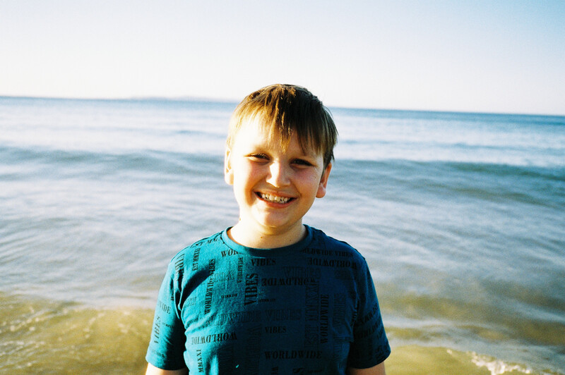Young boy in a blue t-shirt smiling at the camera while standing in shallow ocean water.
