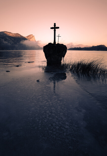 Silhouetted cross on a mound in shallow water at dawn, surrounded by misty landscape.