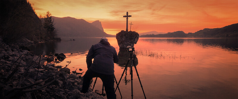 Photographer with tripod-mounted camera silhouetted against a golden sunset over a calm lake with mountains and crosses visible.