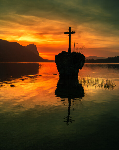 Silhouetted rock formation with crosses reflected in still water at golden hour sunset.