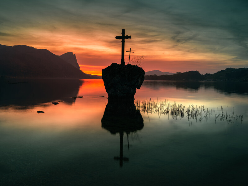 Silhouetted crosses on a structure rising from still water at sunrise, with mountains and dramatic orange sky reflected below.