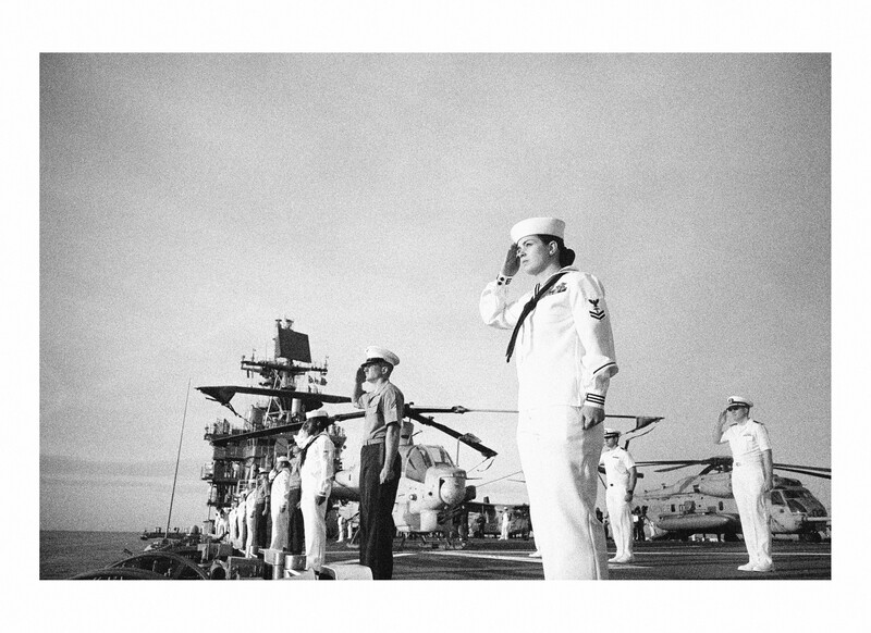 Naval officers in dress whites on an aircraft carrier deck with a helicopter in the background.