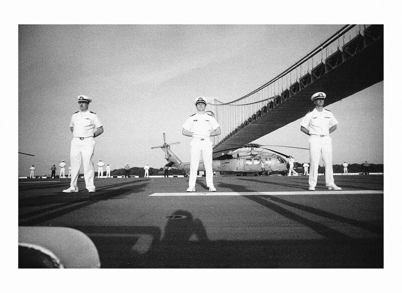 Three naval personnel in white uniforms standing in formation on an aircraft carrier deck beneath a suspension bridge.