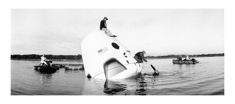 Capsized white aircraft fuselage partially submerged in water with rescue boats nearby.