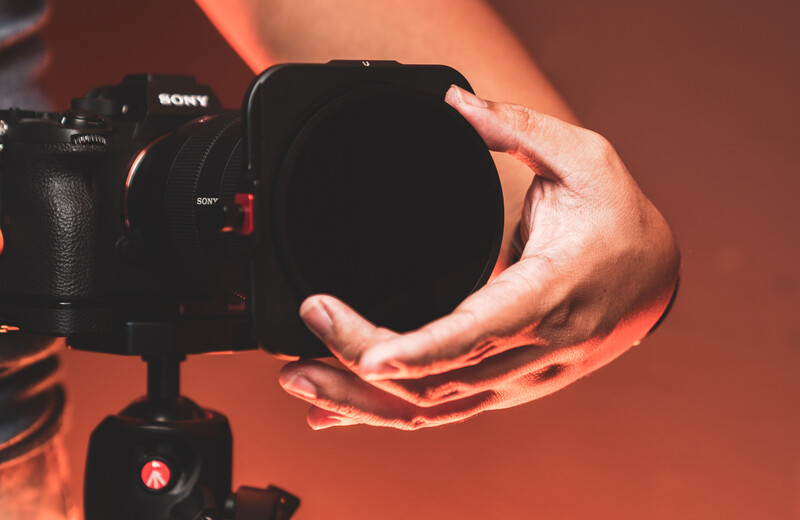 Photographer's hands holding a large lens filter in front of a Sony camera mounted on a gimbal stabilizer.