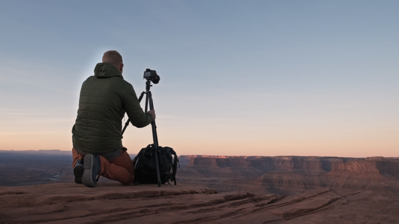 Photographer kneeling on desert terrain while composing a shot with a camera mounted on a tripod during golden hour.