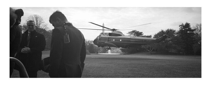 Silhouetted figures observe a helicopter landing on an open grass field.