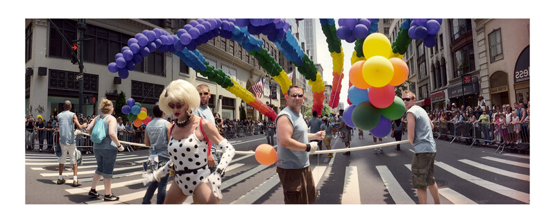 Parade participants holding colorful rainbow balloons and ropes on a city street lined with spectators.