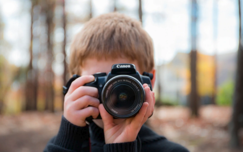 Young child holding a Canon DSLR camera up to their face in an outdoor setting.