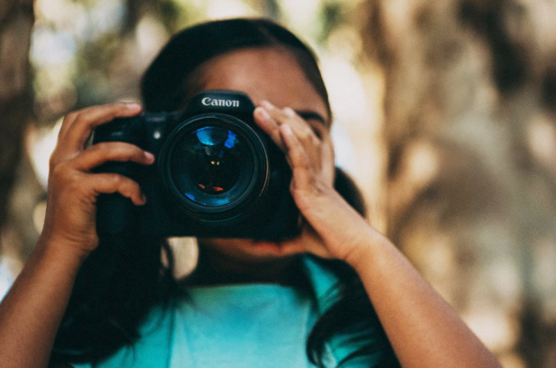 Photographer holding a Canon DSLR camera up to their face, obscuring their identity behind the lens.