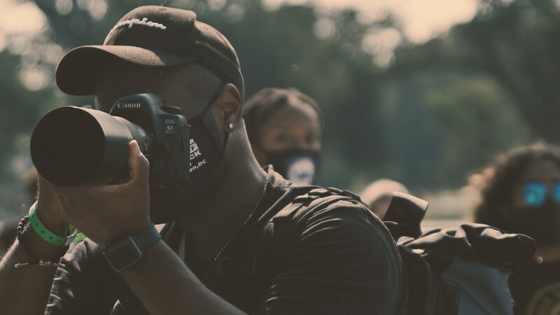 Photographer with Canon DSLR camera and telephoto lens held to face, surrounded by other photographers in outdoor setting.