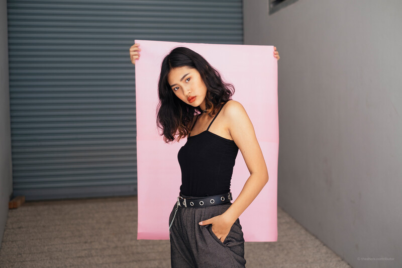 Woman holding pink backdrop in front of her body, wearing black sleeveless top and gray pants with belt.