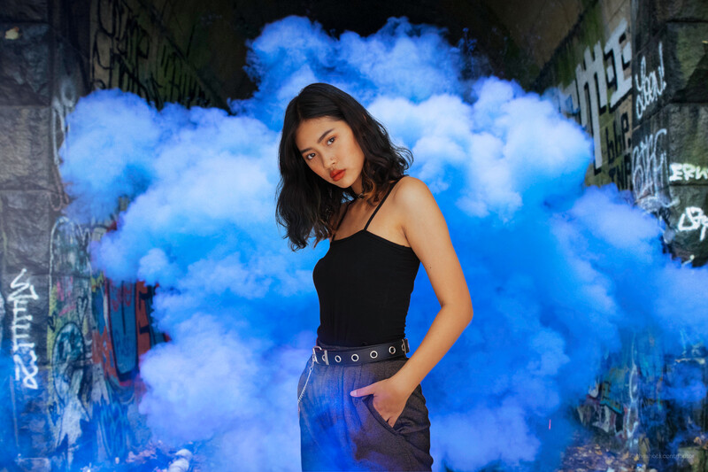 Woman in black tank top and blue pants posing against vibrant blue cloud effect on urban wall.