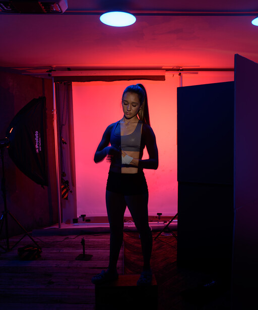 Woman in dark athletic wear posing in front of a glowing pink and red backdrop in a studio setting.