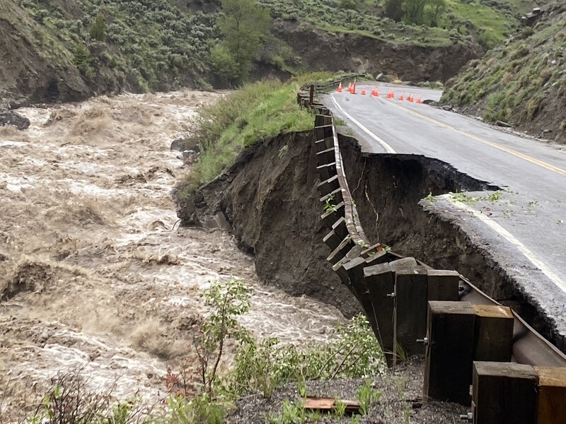 Eroded highway bridge with exposed wooden support structures and churning floodwaters below.