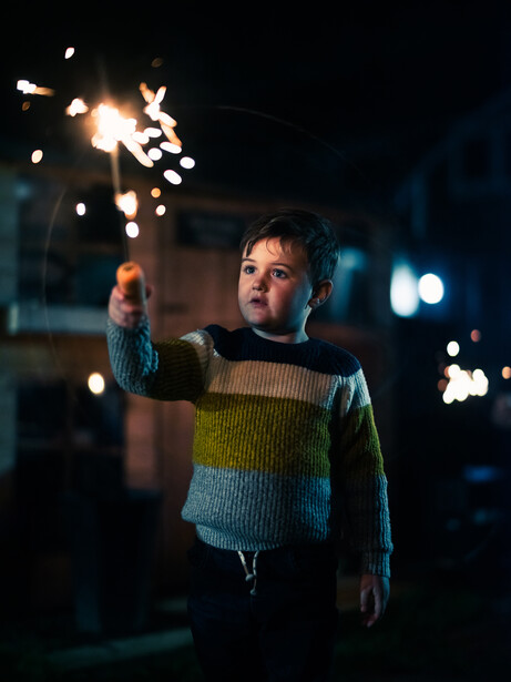 Young child holding a lit sparkler at night, wearing a colorful striped sweater.