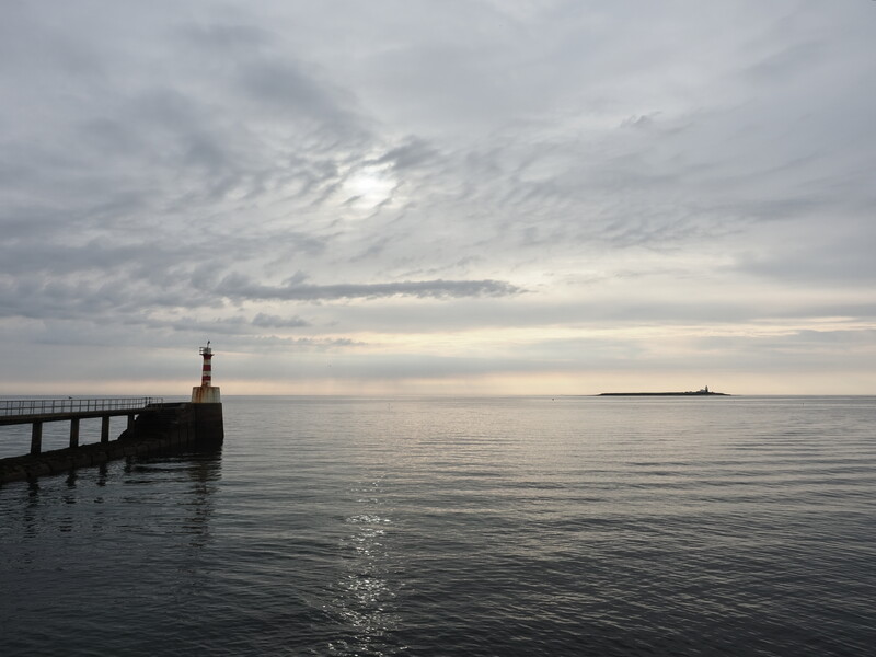 Silhouetted pier and lighthouse at twilight with calm water and overcast sky.