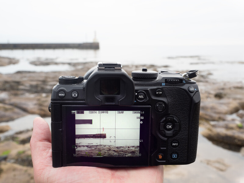 Hand holding a mirrorless camera displaying a histogram on its rear LCD screen, with a rocky coastline blurred in the background.