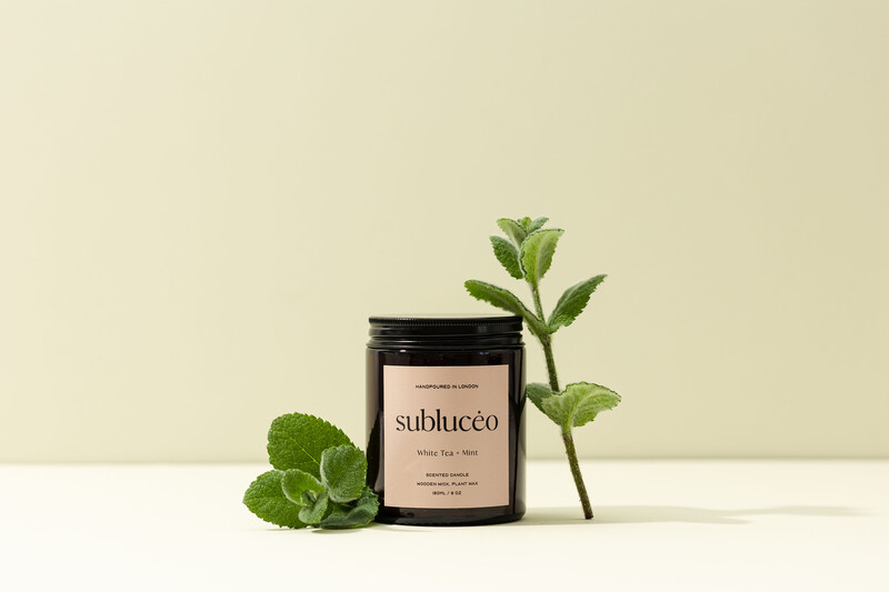 Black glass jar with minimalist label surrounded by fresh mint leaves and a green stem on neutral background.