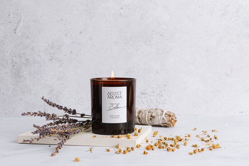 Dark glass candle with white label next to dried lavender stems and scattered chamomile flowers on white surface.