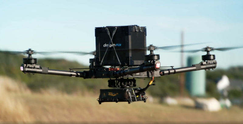 Large industrial drone in flight with extended arms and mounted camera equipment over rural landscape.