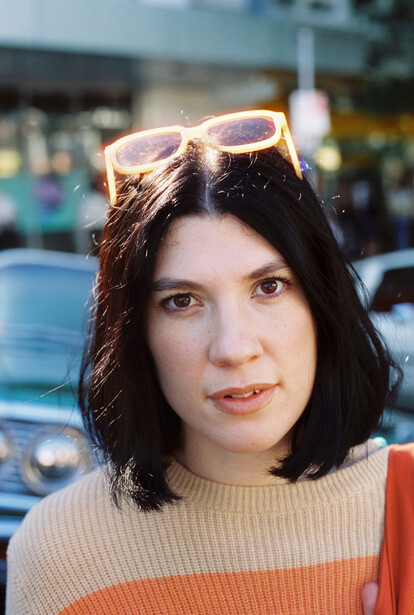 Woman with shoulder-length dark hair wearing a striped knit sweater and sunglasses on her head, outdoors with blurred cars in background.