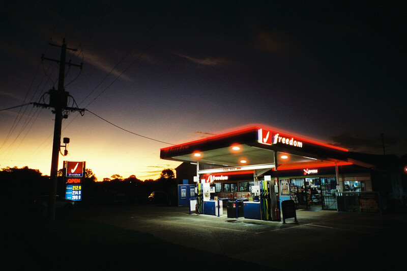 Illuminated gas station with red canopy at dusk, utility pole and warm sky behind it.