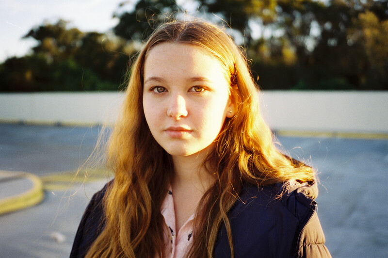 Young woman with long reddish-brown hair looking at camera near a body of water at golden hour.