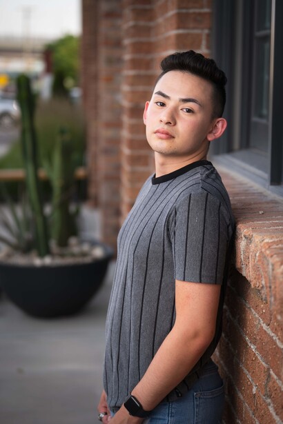 Young man in gray short-sleeve shirt leaning against brick wall, looking toward camera.