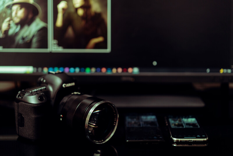 Canon camera with lens on desk beside smartphone, with photo editing software open on monitor behind.