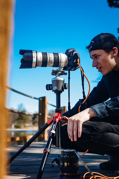 Photographer operating a telephoto lens mounted on a tripod with gimbal head outdoors.