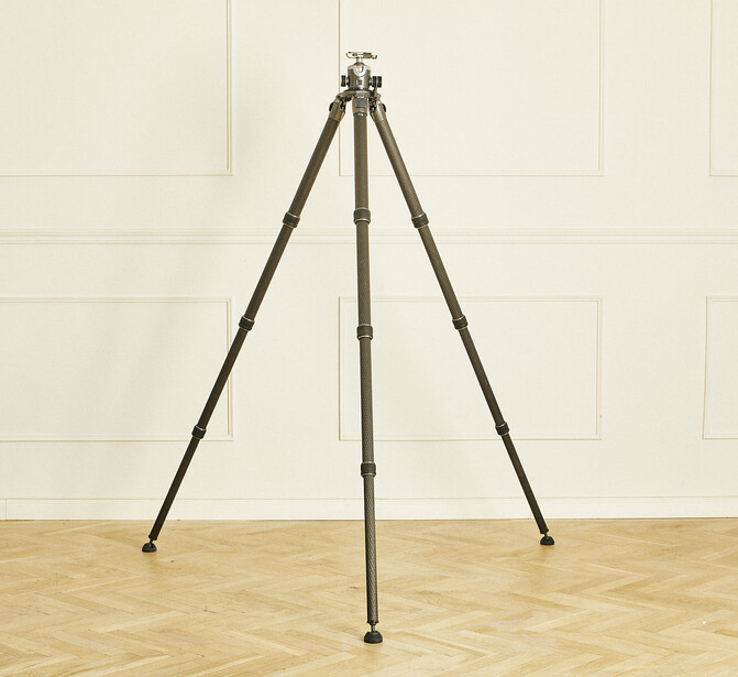 Sturdy tripod with dark brown legs and center column standing on wooden floor against cream-colored wall.