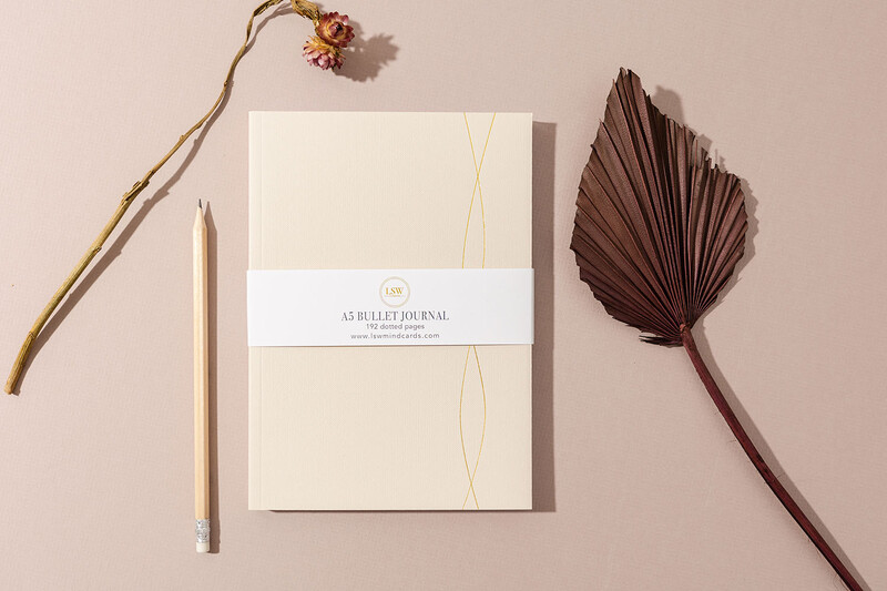 Flat lay composition of a cream-colored bullet journal with gold line detail and band closure, surrounded by dried botanicals and a pencil on a neutral beige background.