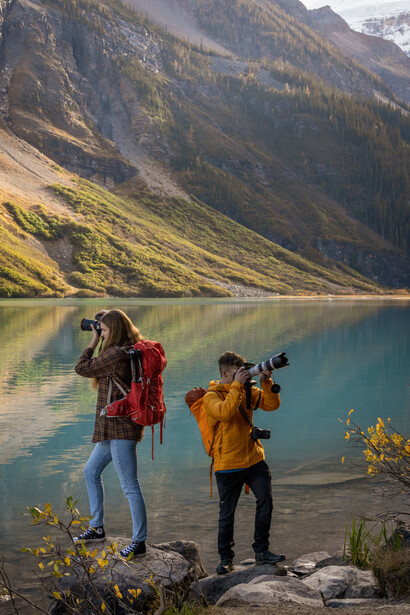 Two photographers with cameras standing on a rocky shore beside a turquoise alpine lake with forested mountains behind.