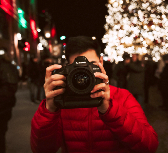 Person in red jacket holding Canon DSLR camera toward viewer at night with blurred holiday lights behind.