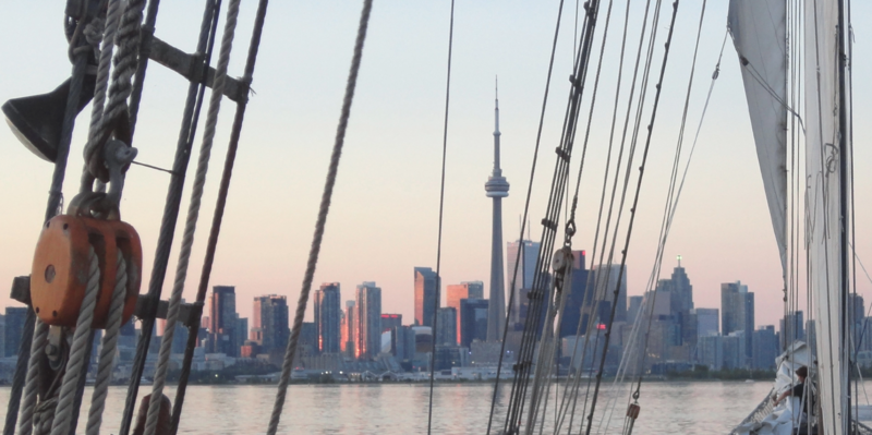View from a sailing vessel toward a city skyline at sunrise, with rigging and pulleys in the foreground.
