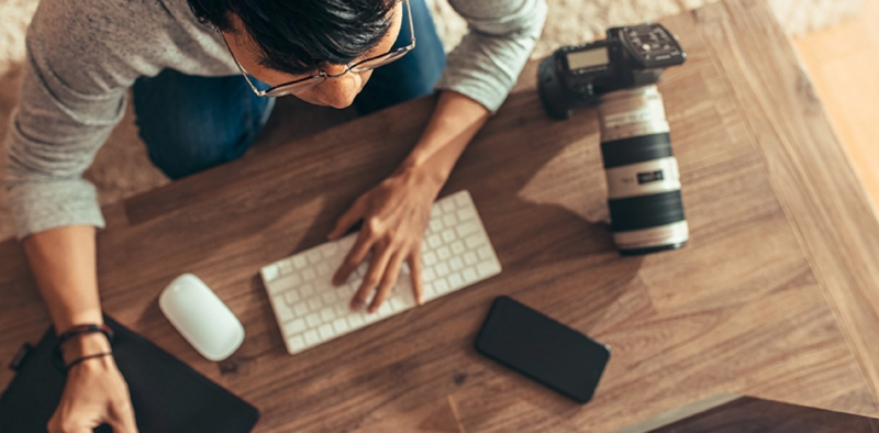 Photographer working at wooden desk with camera, keyboard, mouse, smartphone, and telephoto lens.
