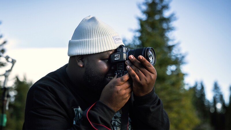 Photographer holding a film camera up to his eye while wearing a white beanie and black jacket outdoors.