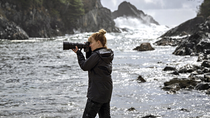 Photographer with telephoto lens standing on rocky coastline with waves and dramatic cliffs.