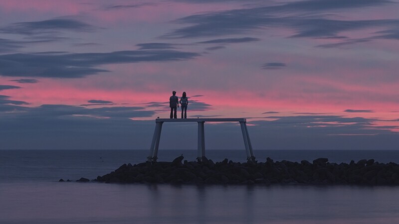 Two silhouetted figures standing on a metal structure in shallow water against a dramatic pink and blue sunset sky.