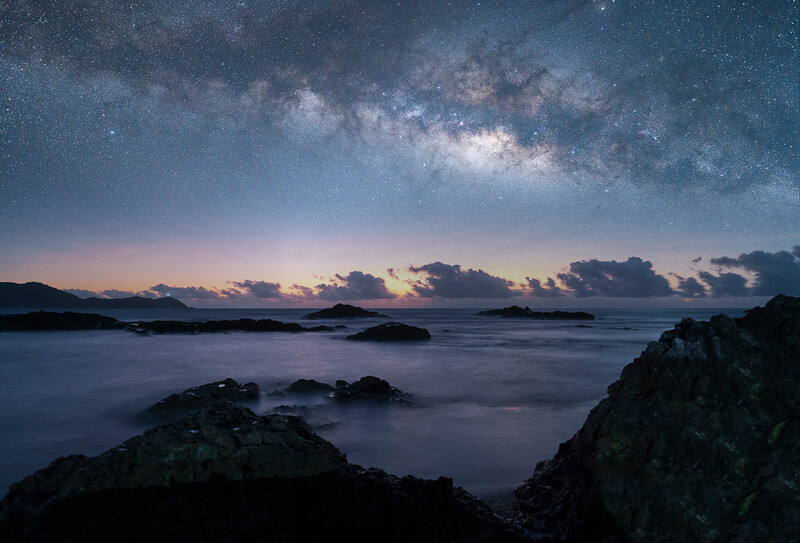 Milky Way galaxy arching over a rocky coastline at twilight, with calm water and islands silhouetted against the horizon.