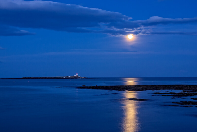 Full moon illuminating calm ocean waters at twilight, with distant ship and rocky shoreline.